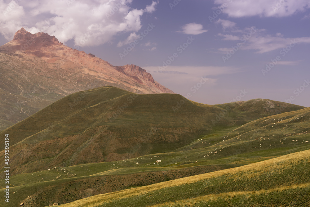 Mountain landscape - grand orange pink bizarre rock of canyon in bright sunny summer day with fluffy white cloud in blue sky, velvety hilly slopes with lush green meadow. Amazing trip in Dagestan.