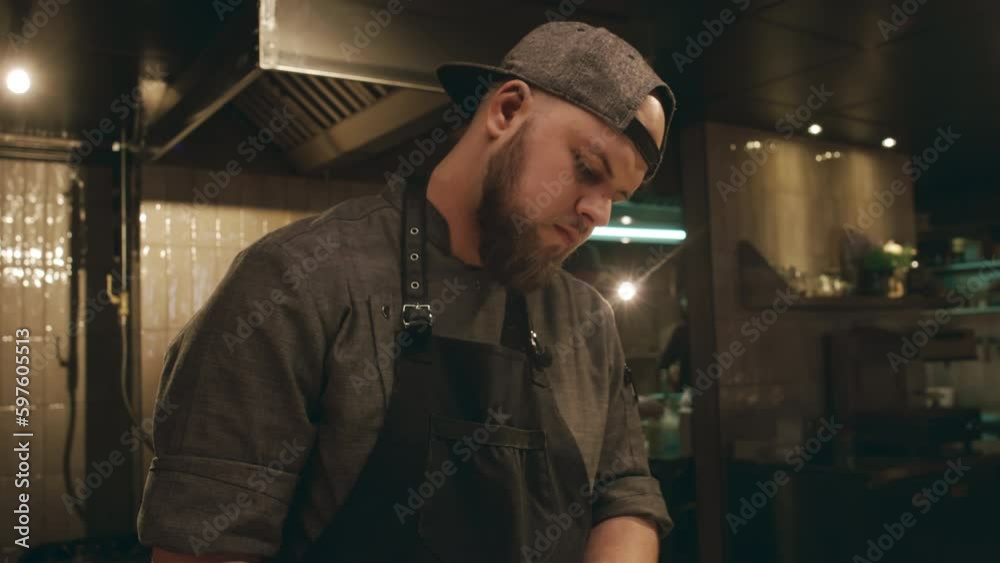 Medium tilting shot of face and hands of busy young Caucasian man in chef uniform, cap and gloves working in restaurant kitchen and slicing ball of mozzarella cheese with knife on chopping board Medium tilting shot of face and hands of busy young Caucasian man in chef uniform, cap and gloves working in restaurant kitchen and slicing ball of mozzarella cheese with knife on chopping board