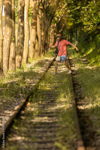 Backpacker man, walking along train tracks, observing Portugal's lush green forest in summertime. Wearing hat, pink t-shirt and short with sandals. Sunbeams, vertical