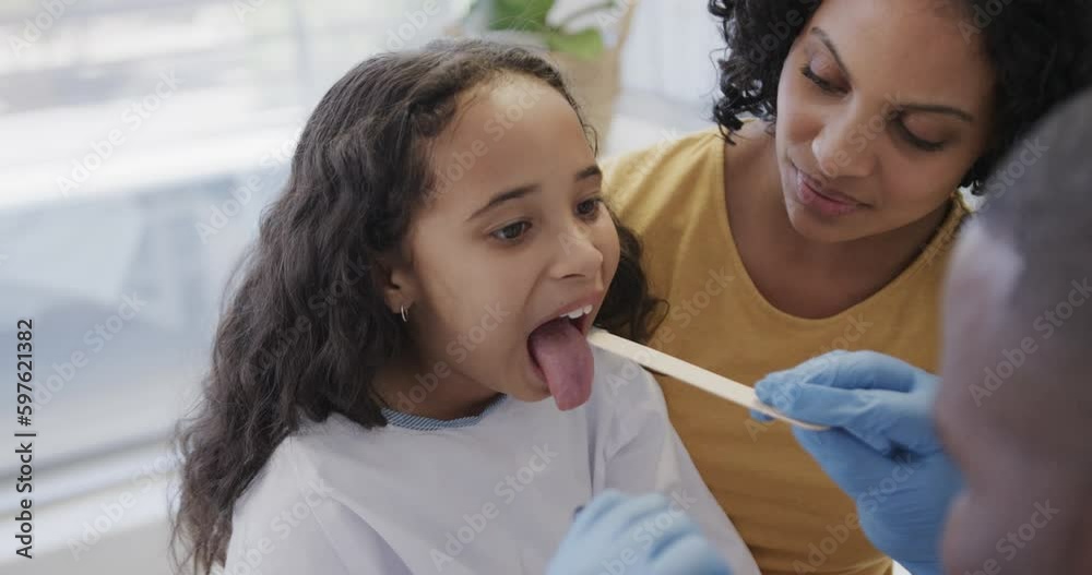 Happy diverse doctor checking sick girl patient's throat with mother in hospital in slow motion