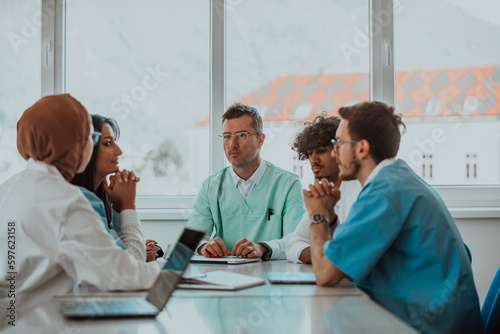 A group of multiethnic medical professionals including doctors, surgeons, and nurses are gathered in a hospital setting discussing patient care and using modern technology to address challenges in photo