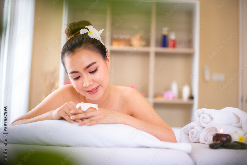 Naklejka premium Beautiful asian woman lying on a comfortable white bed in a peaceful spa room while holding white frangipani flowers.