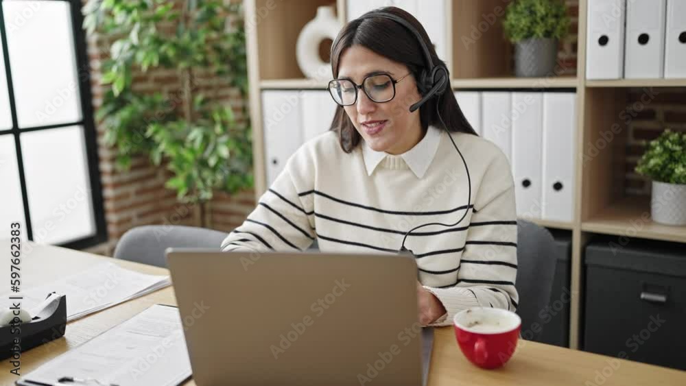 Young beautiful hispanic woman call center agent having video call writing on document at office