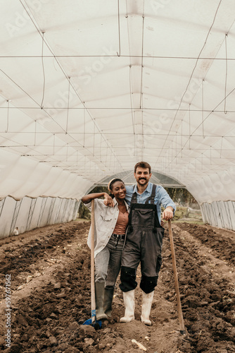 Portrait of a multiracial couple standing in a work outfit and preparing va soil in a greenhouse.