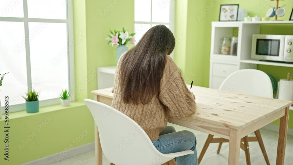 Young beautiful hispanic woman writing on notebook tired stretching arms at kitchen Young beautiful hispanic woman writing on notebook tired stretching arms at kitchen