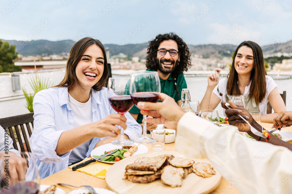 Happy diverse group of friends enjoying dinner barbecue in summer at home terrace. Two young people cheering with red wine at bbq party. Food and family moments concept Happy diverse group of friends enjoying dinner barbecue in summer at home terrace. Two young people cheering with red wine at bbq party. Food and family moments concept