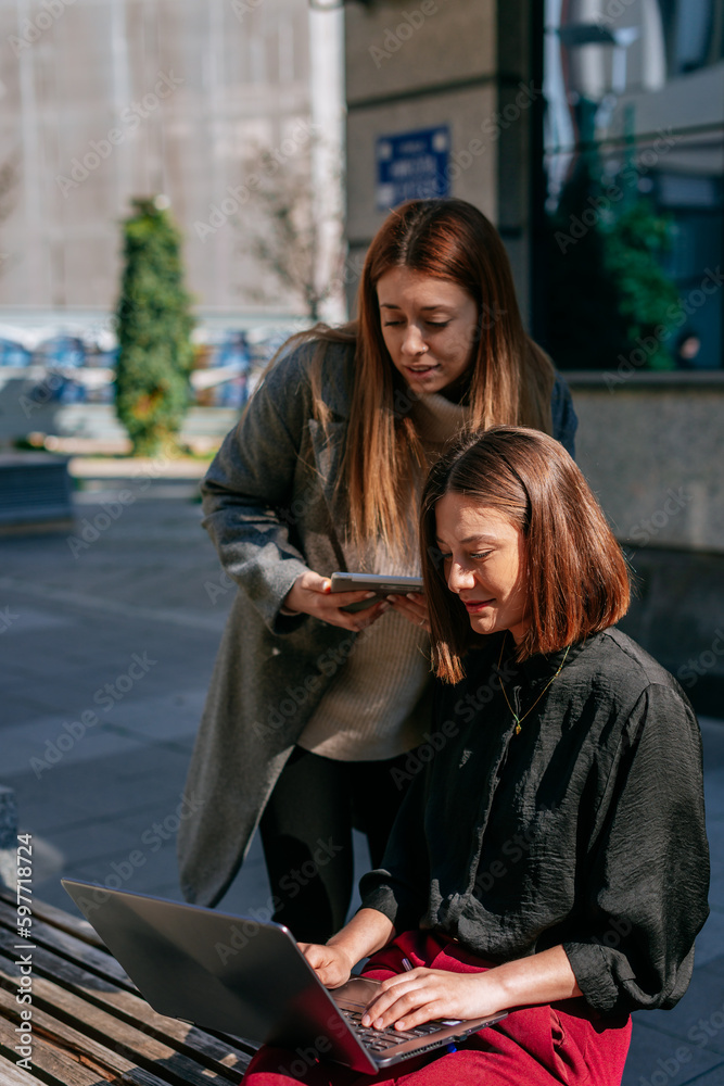 A shot of two young, attractive businesswomen working together out in the city. Brunette businesswoman sitting on a bench and working on the lap top while her blonde colleague is standing behind her A shot of two young, attractive businesswomen working together out in the city. Brunette businesswoman sitting on a bench and working on the lap top while her blonde colleague is standing behind her