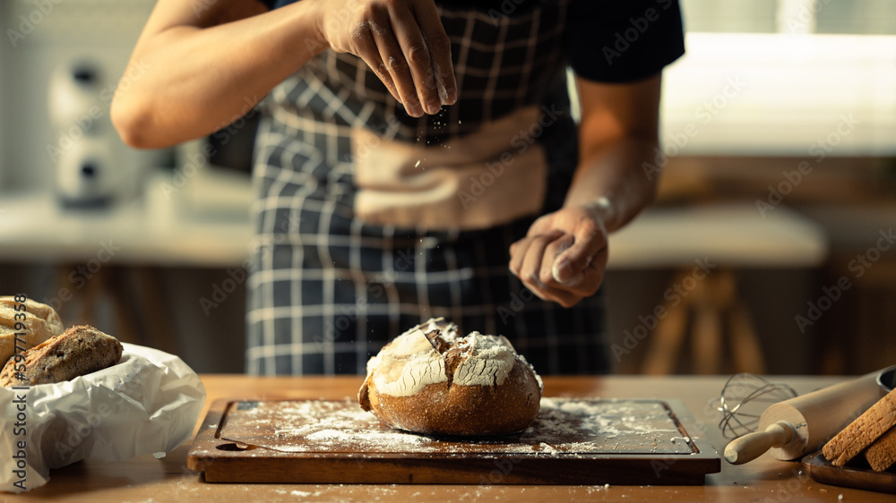 Male baker wearing apron sifting flour on dough, making bread in kitchen