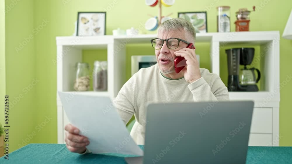 Middle age grey-haired man talking on smartphone reading document at home Middle age grey-haired man talking on smartphone reading document at home