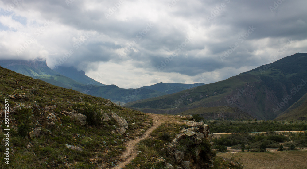 Naklejka premium Dramatic landscape - the picturesque Baksan valley with a country road among the hills against the background of cloudy sky and foggy haze in the Elbrus region in the North Caucasus and copy space