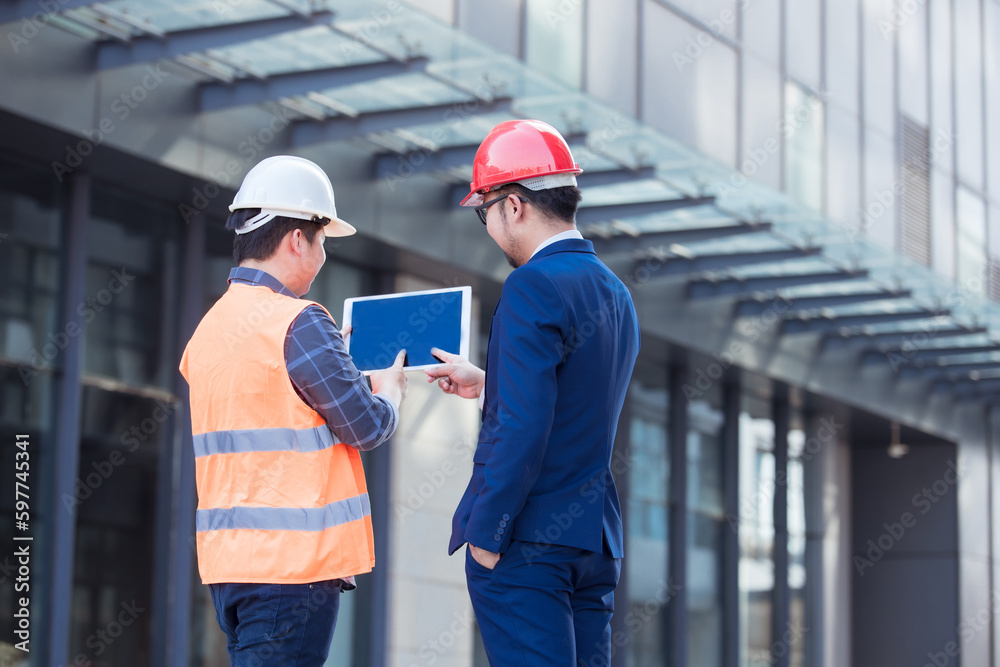 Two Industry Engineers Stand in modern building construction, Use Digital Tablet, Have Discussion. Engineers wearing safety vest and hard hat. 