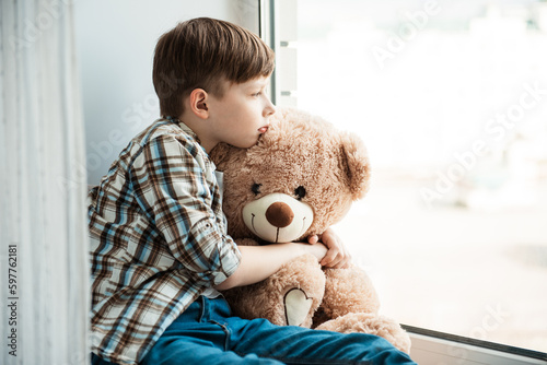 Sadness outside the window. One child boy abandoned in an old abandoned house with a teddy bear. He is his only friend