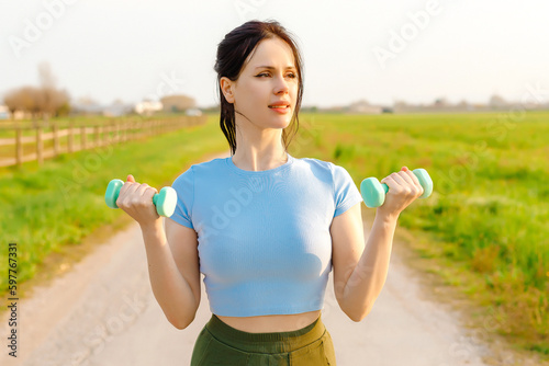 Smiling girl doing sports outdoors with dumbbells lifting weights.