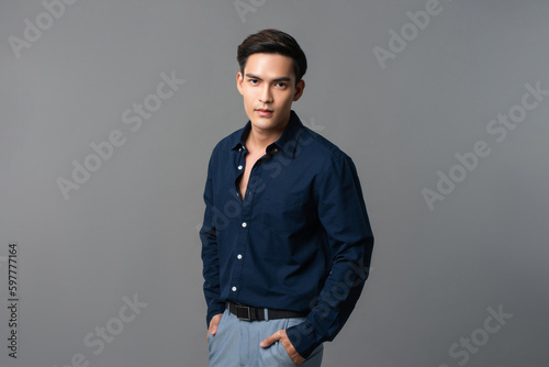 Studio portrait of young handsome Asian man wearing dark blue shirt standing in light gray isolated background with hands in pockets