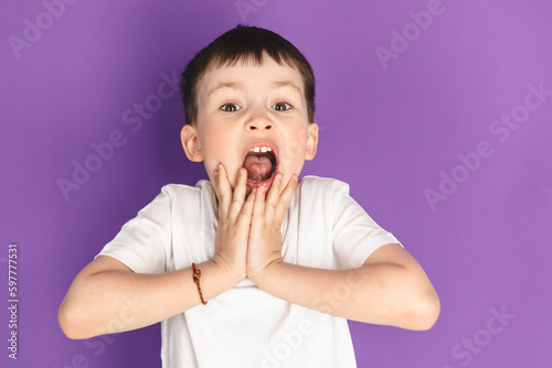 Oh my god  wow  Portrait of funny amazed preschool boy looking at camera with shocked astonished expression and keeping hands on face  screaming in surprise studio shot isolated on purple background.