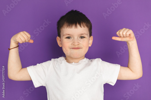 Little brunette boy in white t-shirt show strong arm gesture, powerful isolated purple background. Funny little power super hero kid showing muscles. Strength, confidence or defense from bullying.