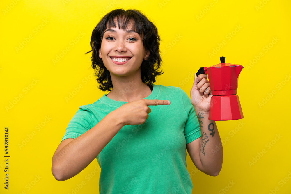 Young Argentinian woman holding coffee pot isolated on yellow background and pointing it Young Argentinian woman holding coffee pot isolated on yellow background and pointing it