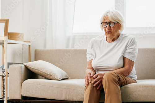 Elderly woman severe pain in her leg sitting on the couch, health problems in old age, poor quality of life. Grandmother with gray hair holds on to her sore knee, problems with joints and ligaments. © SHOTPRIME STUDIO