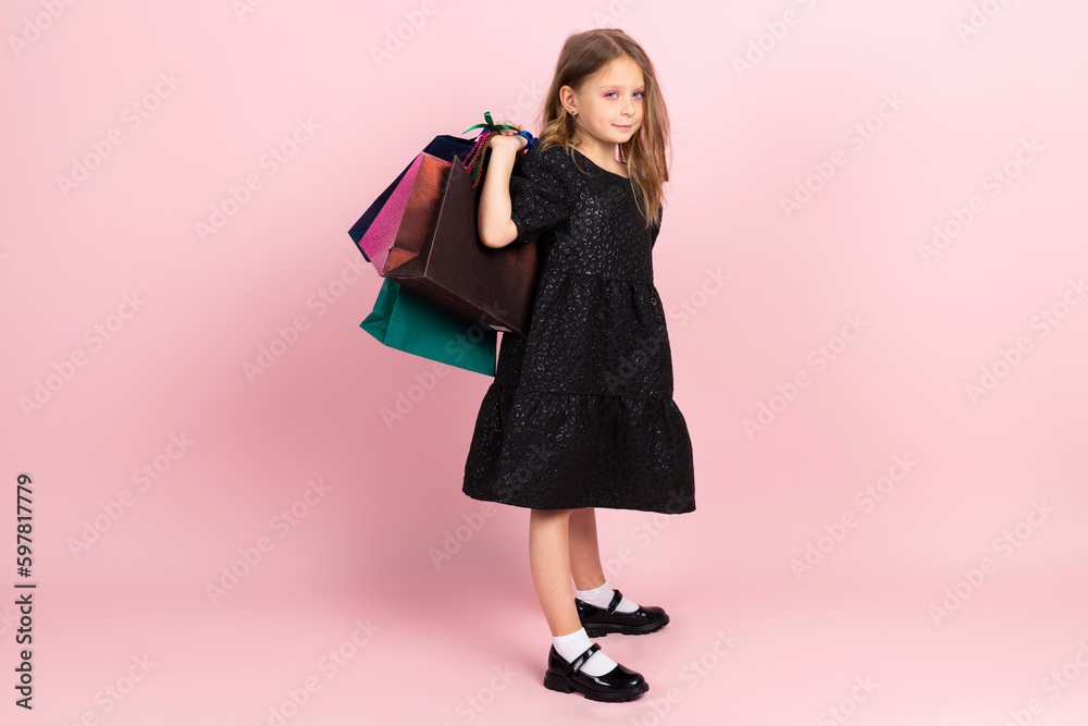 Child shopping. Charming little girl in black stylish summer dress and slight pink makeup, posing with colorful shopping bags. Isolated on pink background. Child shopping. Charming little girl in black stylish summer dress and slight pink makeup, posing with colorful shopping bags. Isolated on pink background.