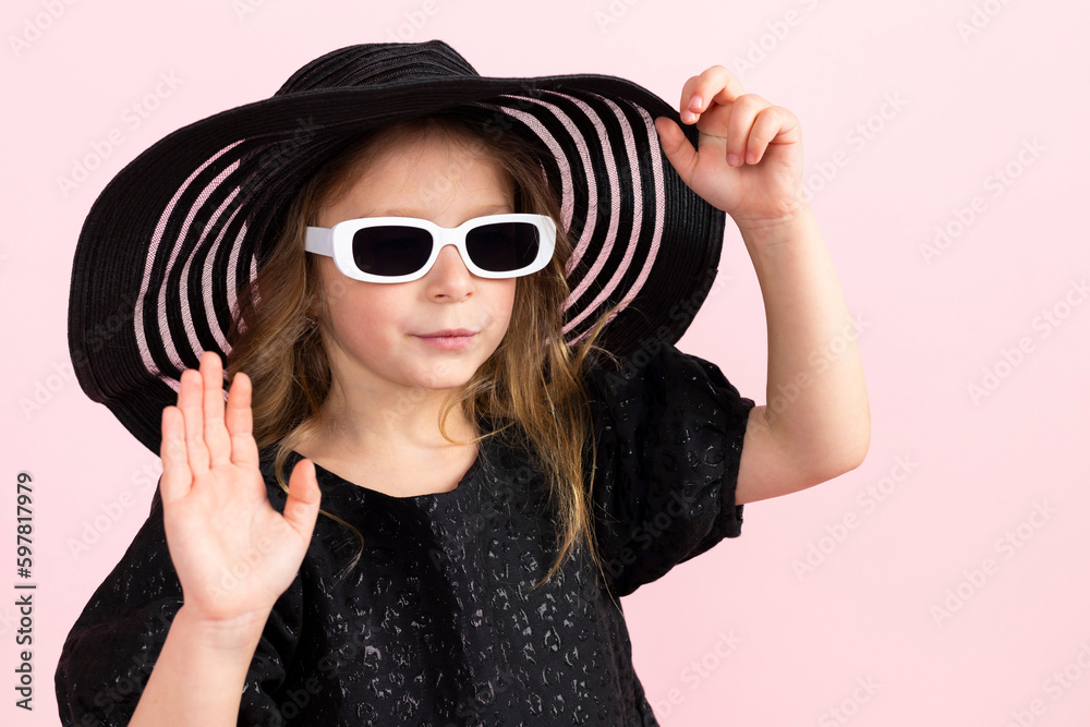 Charming little girl in black stylish summer dress, sun hat and a cool white sunglasses. Isolated on pink background.