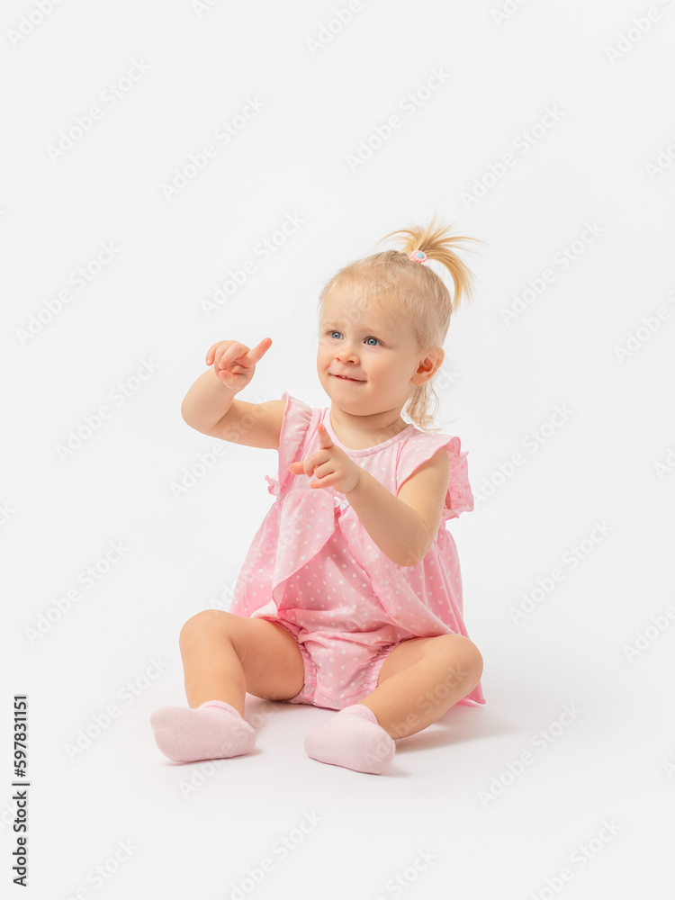A 1-2-year-old Caucasian girl in a pink dress is sitting on the floor looking and pointing with her fingers on a white background. Copy space. A 1-2-year-old Caucasian girl in a pink dress is sitting on the floor looking and pointing with her fingers on a white background. Copy space.