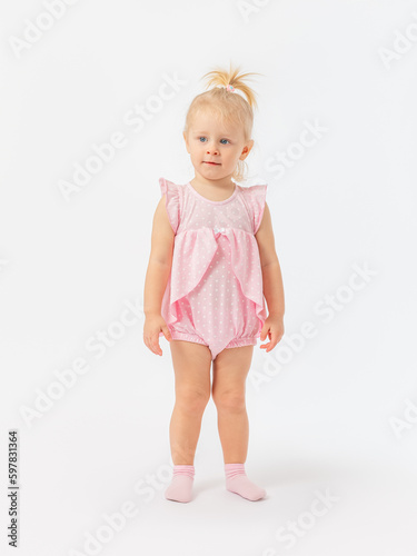 A 2-year-old baby in a pink dress stands calmly and attentively looks at something, studies, listens on a white background.