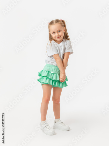 A 5-year-old girl with long hair in a T-shirt and a green skirt stands with her shoulders tucked up and her arms crossed in sneakers on a white background. Looking into the camera