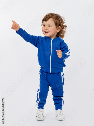 A cheerful little boy of 1-2 years old stands, smiles and points his finger up in a blue tracksuit and sneakers on a white background.