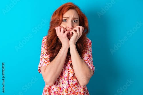 Anxiety - Caucasian redhead woman wearing floral dress over blue background covering his mouth with hands scared from something or someone bitting nails.