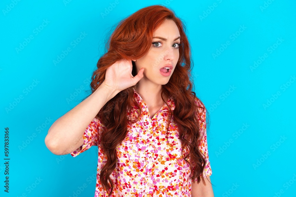 Oh my God! Funny astonished Caucasian redhead woman wearing floral dress over blue background opening mouth widely looking aside, with hand near ear trying to listen to gossips. Oh my God! Funny astonished Caucasian redhead woman wearing floral dress over blue background opening mouth widely looking aside, with hand near ear trying to listen to gossips.