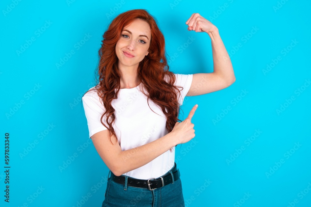 Smiling young redhead woman wearing white T-shirt over blue background raises hand to show muscles, feels confident in victory, strong and independent. Smiling young redhead woman wearing white T-shirt over blue background raises hand to show muscles, feels confident in victory, strong and independent.