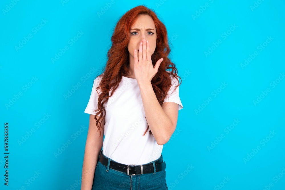 Oh! I think I said it! Close up portrait young redhead woman wearing white T-shirt over blue background cover open mouth by hand palm, look at camera with big eyes.