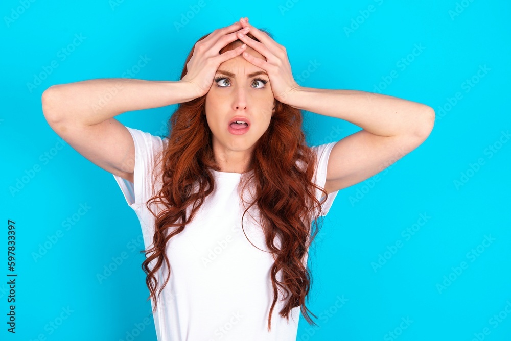 Horrible, stress, shock. Portrait emotional crazy young redhead woman wearing white T-shirt over blue background clasping head in hands. Emotions, facial expression concept. Horrible, stress, shock. Portrait emotional crazy young redhead woman wearing white T-shirt over blue background clasping head in hands. Emotions, facial expression concept.