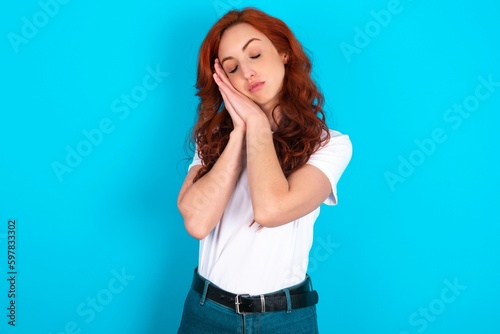 Relax and sleep time. Tired young redhead woman wearing white T-shirt over blue background with closed eyes leaning on palms making sleeping gesture.