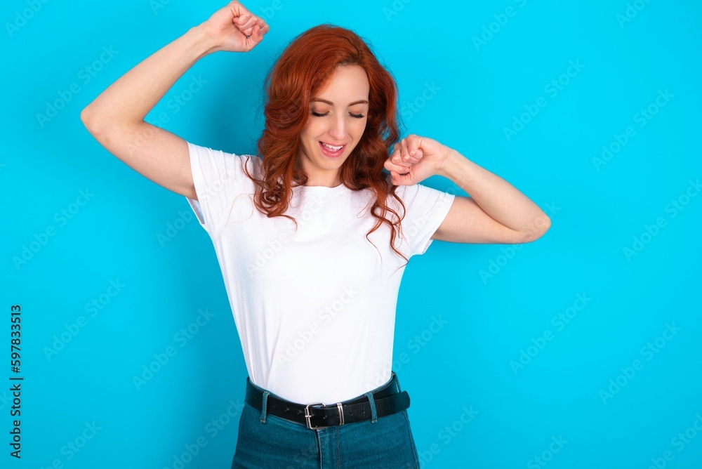 Attractive young redhead woman wearing white T-shirt over blue background celebrating a victory punching the air with his fists and a beaming toothy smile. Attractive young redhead woman wearing white T-shirt over blue background celebrating a victory punching the air with his fists and a beaming toothy smile.