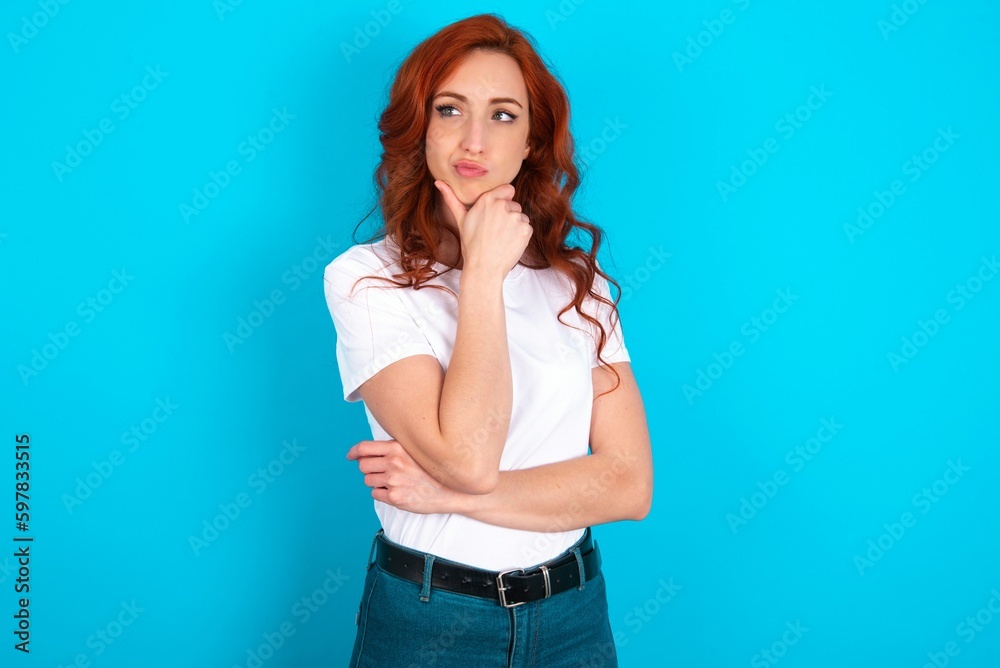 young redhead woman wearing white T-shirt over blue background with hand under chin and looking sideways with doubtful and skeptical expression, suspect and doubt. young redhead woman wearing white T-shirt over blue background with hand under chin and looking sideways with doubtful and skeptical expression, suspect and doubt.