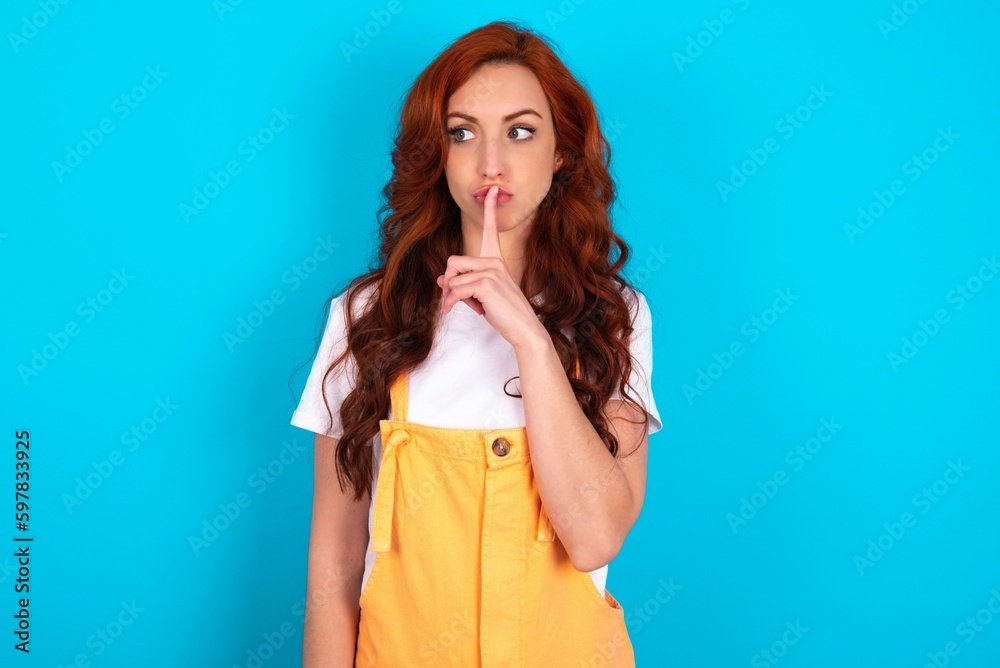 Young redhead woman wearing orange overall over blue background makes silence gesture, keeps index finger to lips makes hush sign. Asks not to share secret. Young redhead woman wearing orange overall over blue background makes silence gesture, keeps index finger to lips makes hush sign. Asks not to share secret.