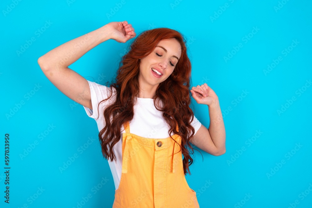 Photo of upbeat Young redhead woman wearing orange overall over blue background has fun and dances carefree wear being in perfect mood makes movements. Spends free time on disco party Photo of upbeat Young redhead woman wearing orange overall over blue background has fun and dances carefree wear being in perfect mood makes movements. Spends free time on disco party