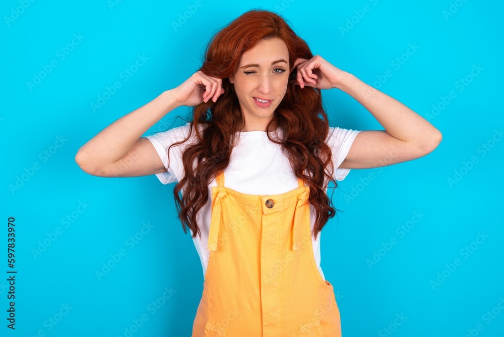 Happy Young redhead woman wearing orange overall over blue background ignores loud music and plugs ears with fingers asks to turn off sound Happy Young redhead woman wearing orange overall over blue background ignores loud music and plugs ears with fingers asks to turn off sound