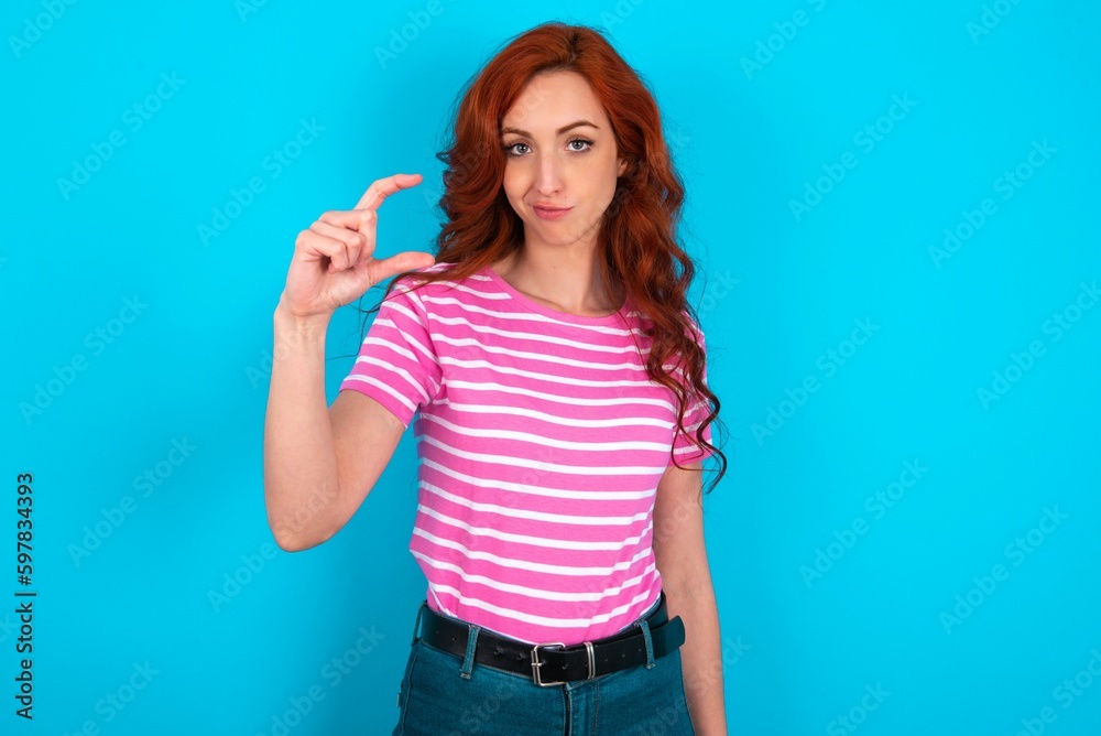 Displeased young redhead woman wearing striped T-shirt over blue background shapes little hand sign demonstrates something not very big. Body language concept. Displeased young redhead woman wearing striped T-shirt over blue background shapes little hand sign demonstrates something not very big. Body language concept.