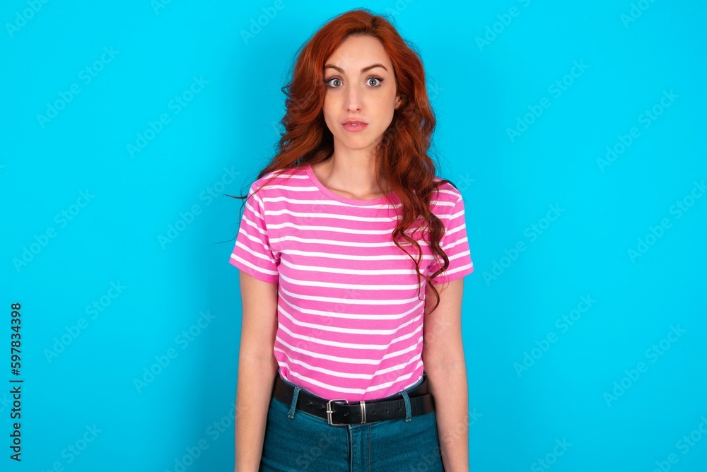 Stunned young redhead woman wearing striped T-shirt over blue background stares reacts on shocking news. Astonished woman holds breath Stunned young redhead woman wearing striped T-shirt over blue background stares reacts on shocking news. Astonished woman holds breath