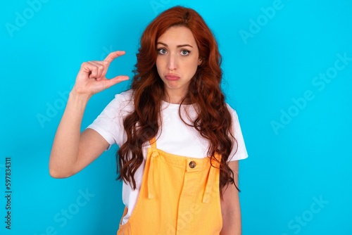 Young redhead woman wearing orange overall over blue background purses lip and gestures with hand, shows something very little.
