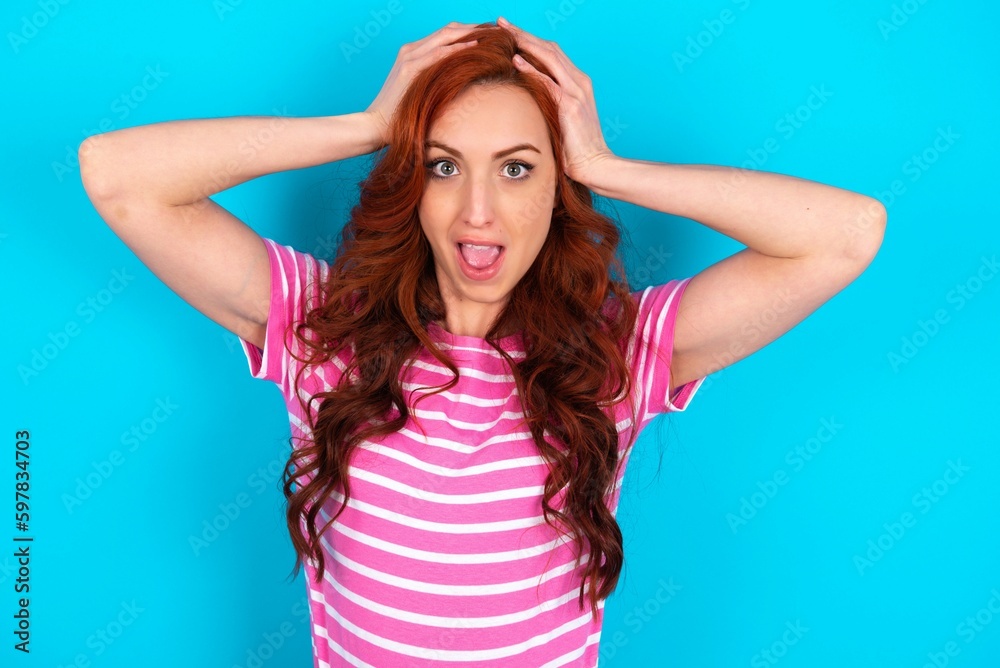 Cheerful overjoyed young redhead woman wearing striped T-shirt over blue background reacts rising hands over head after receiving great news.