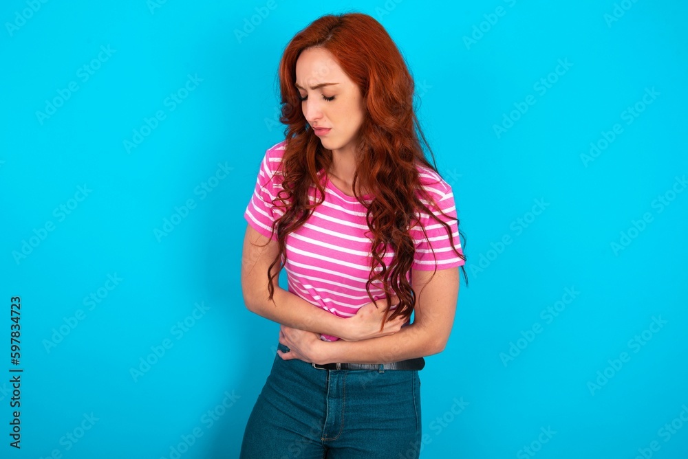 young redhead woman wearing striped T-shirt over blue background got stomachache young redhead woman wearing striped T-shirt over blue background got stomachache