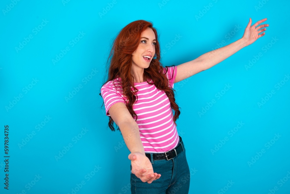 Funny astonished young redhead woman wearing striped T-shirt over blue background look empty space with arms opened ready to catch something. Funny astonished young redhead woman wearing striped T-shirt over blue background look empty space with arms opened ready to catch something.
