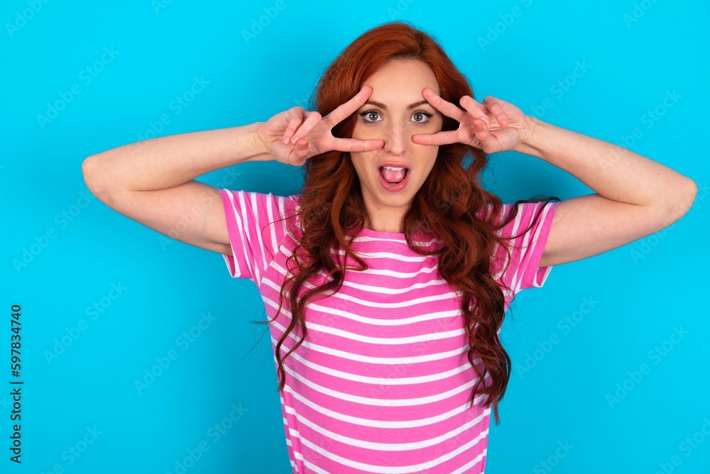 Cheerful positive young redhead woman wearing striped T-shirt over blue background shows v-sign near eyes open mouth Cheerful positive young redhead woman wearing striped T-shirt over blue background shows v-sign near eyes open mouth