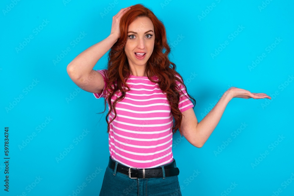 Shocked amazed surprised young redhead woman wearing striped T-shirt over blue background hold hand offering proposition Shocked amazed surprised young redhead woman wearing striped T-shirt over blue background hold hand offering proposition