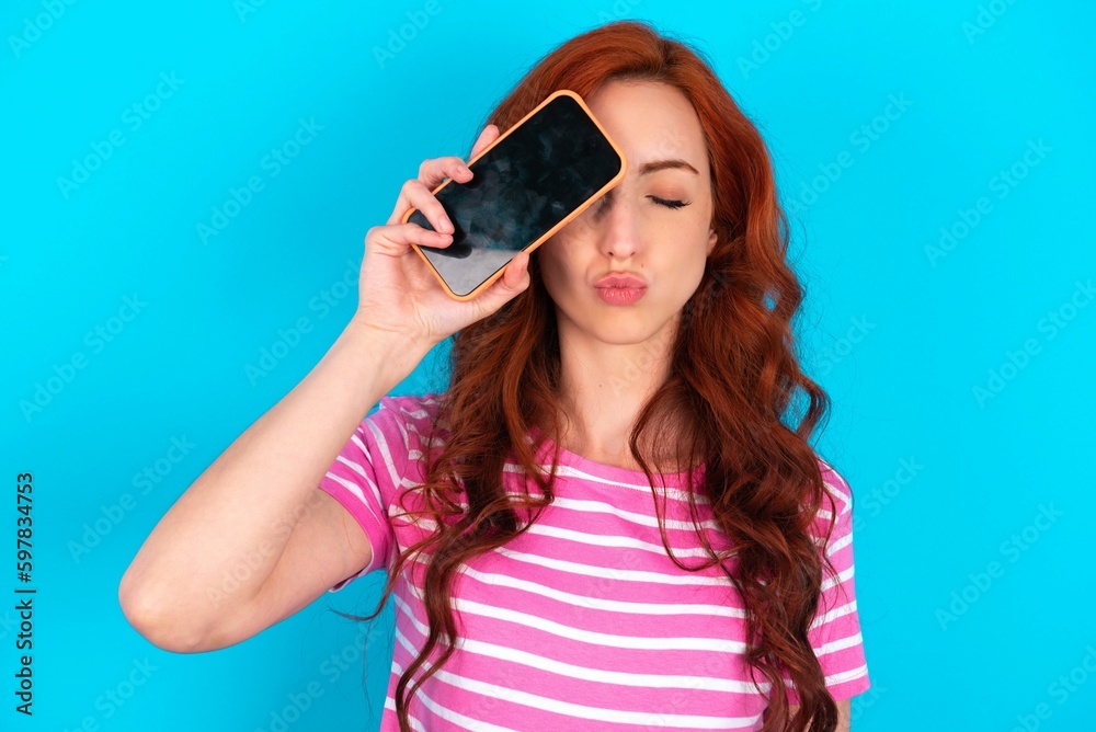 Adorable young redhead woman wearing striped T-shirt over blue background holding modern device covering eye with lips pouted Adorable young redhead woman wearing striped T-shirt over blue background holding modern device covering eye with lips pouted