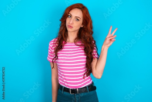 young redhead woman wearing striped T-shirt over blue background makes peace gesture keeps lips folded shows v sign. Body language concept © Jihan