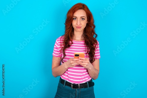 Portrait of serious confident young redhead woman wearing striped T-shirt over blue background holding phone in two hands